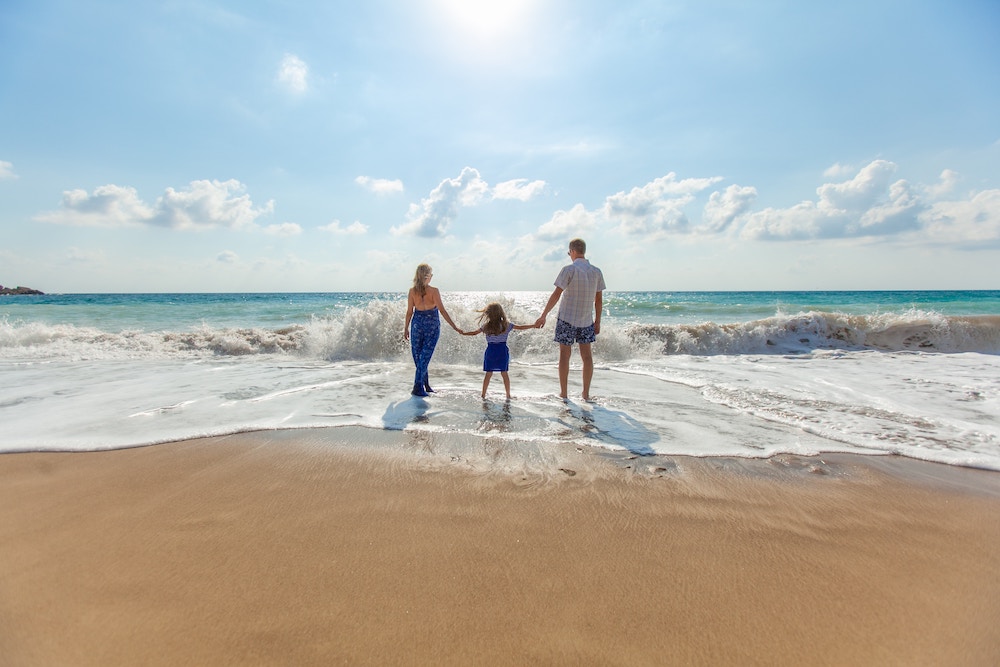 A mother and father holding their daughters hand as they stand on the beach with the waves crashing around them.