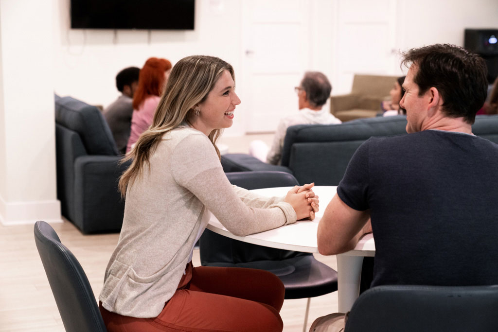A young woman and man smiling and sharing a conversation with more people sitting on a couch in the background.