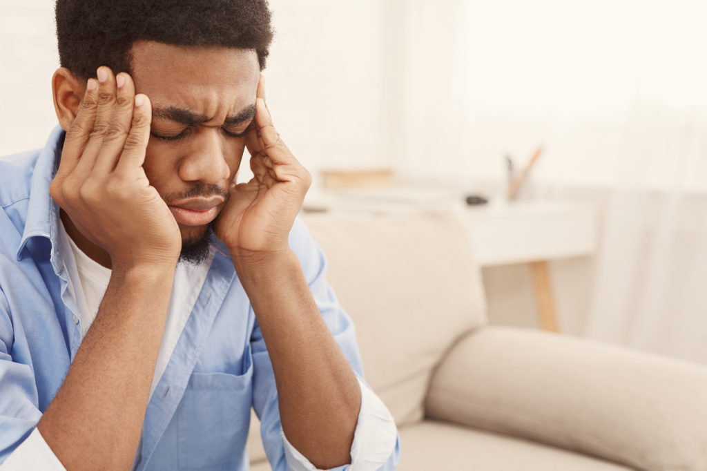 An african-american male sitting on the couch rubbing his temple, he is in pain.
