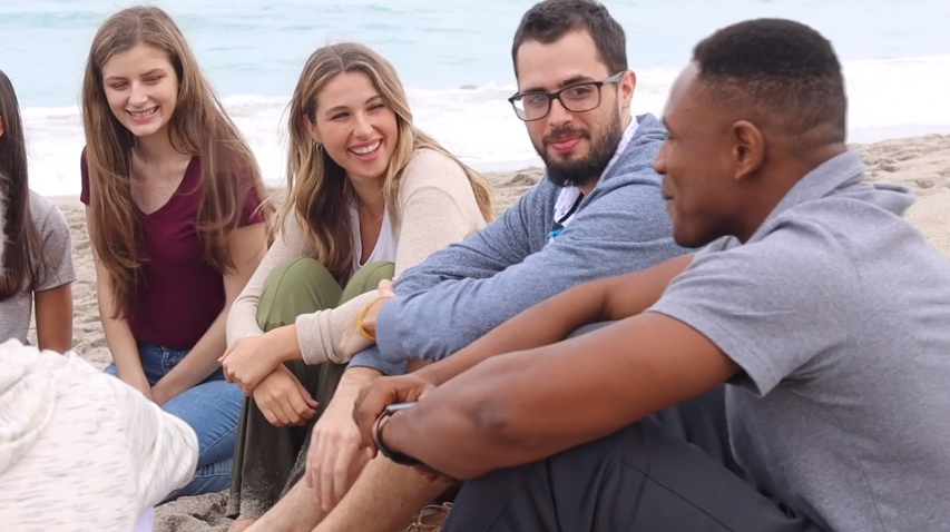 Group of men and women smiling on a beach during a group therapy session.