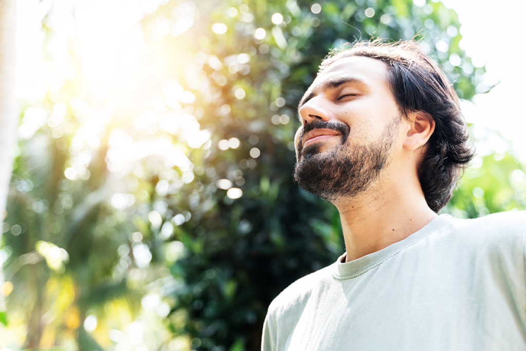 A young man with a beard is smiling in the sun with his eyes closed. He looks at peace.