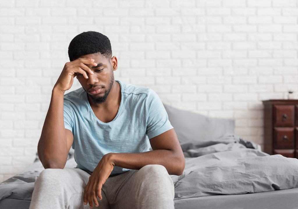 An african-american male sitting on the edge of an unmade bed, looking troubled.