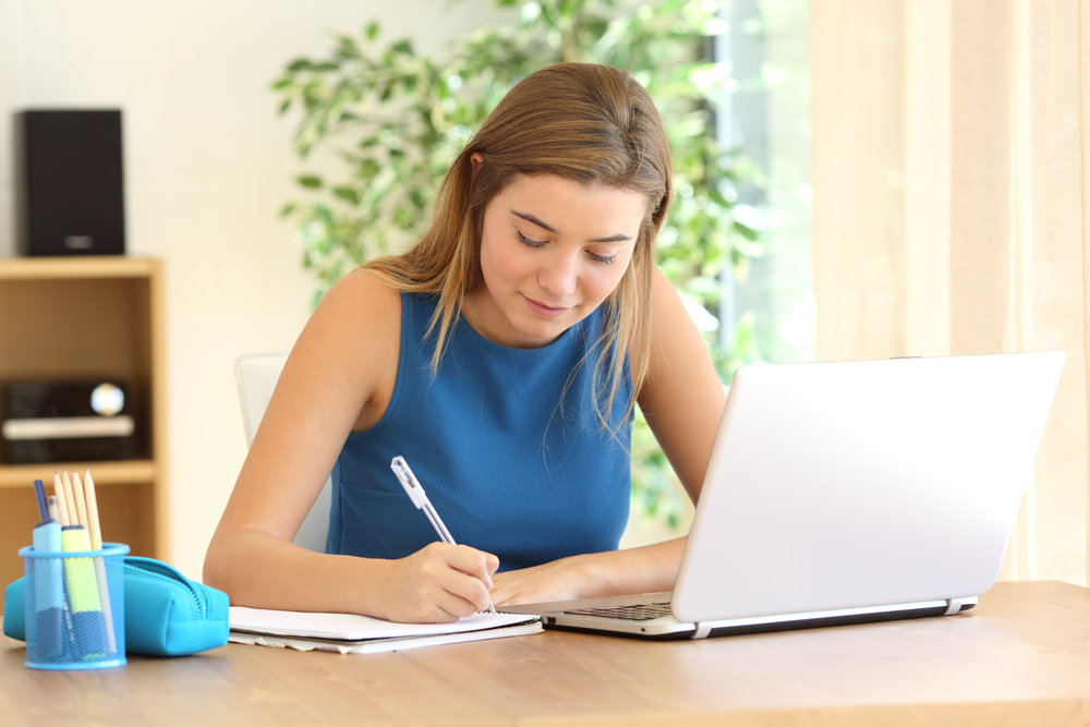 A caucasian woman writing on a notepad as she sits at her desk with her laptop in front of her.