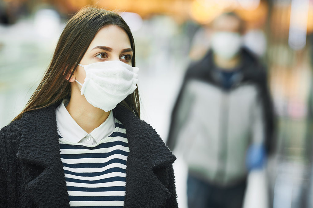 A brunette woman walking in the streets while wearing a mask, there is a man wearing a mask in the background behind her.