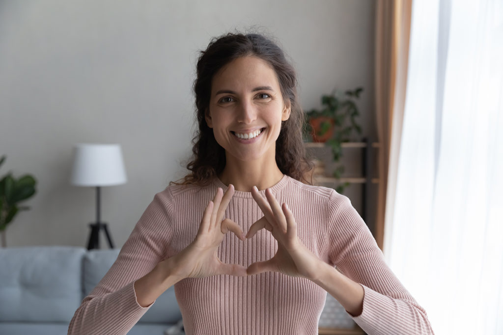 A brunette woman smiling at the camera making a heart shape with her hands.