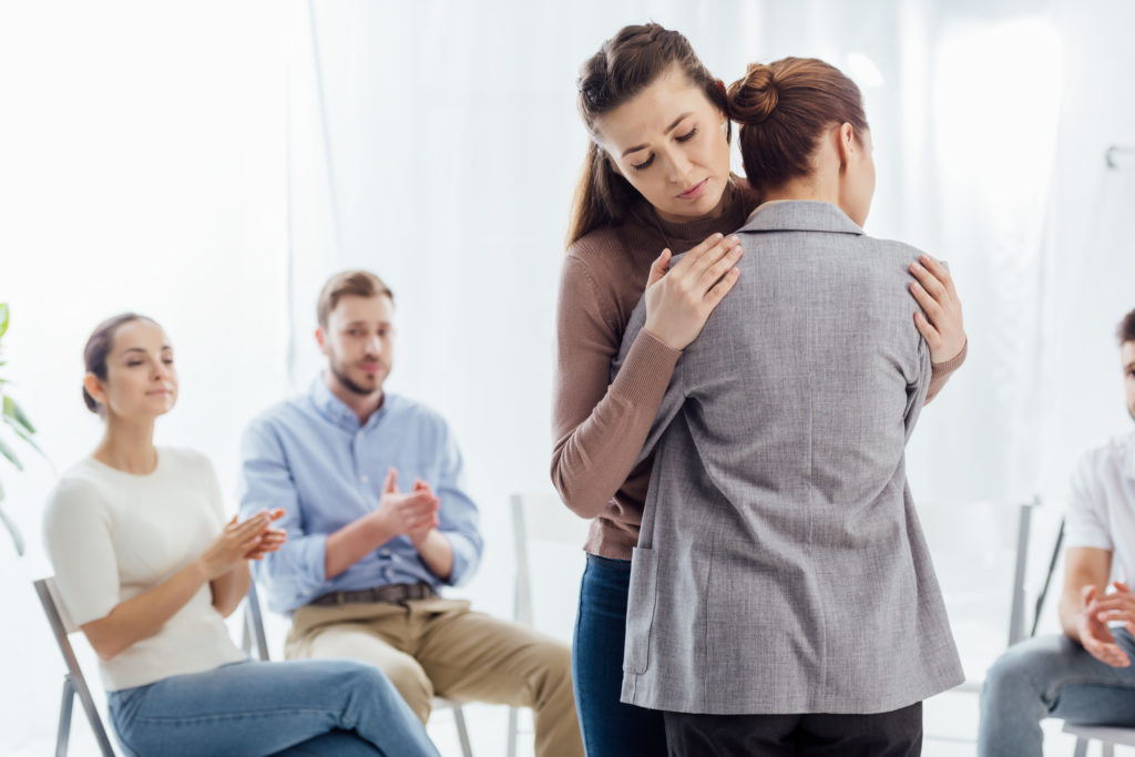 Two caucasian women sharing a hug during a group therapy session, the other patients are clapping in the background.