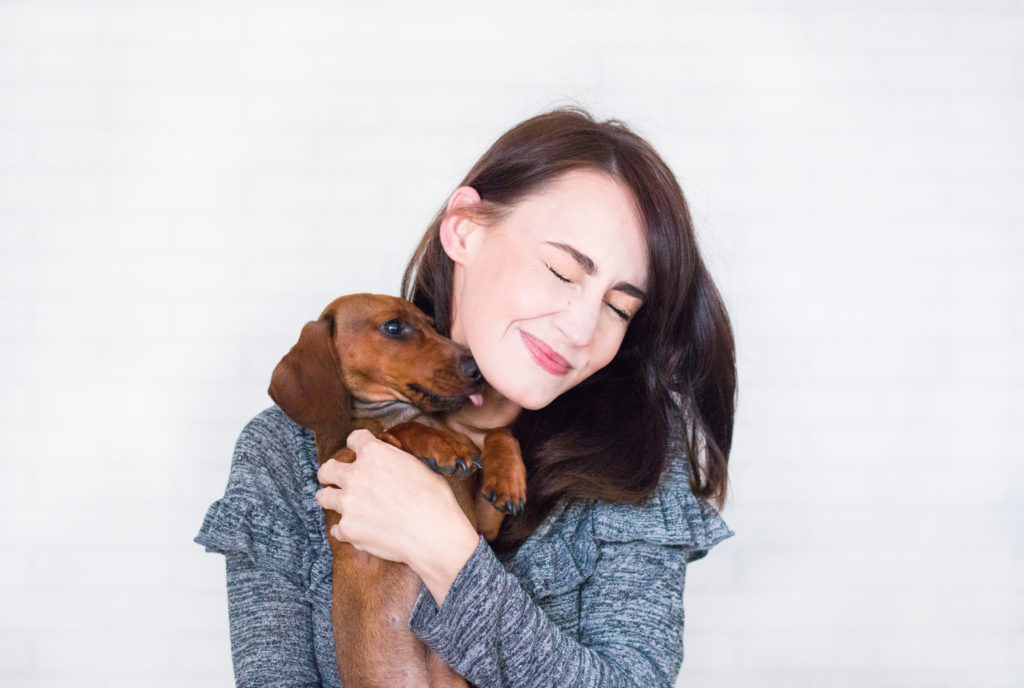 A brunette woman holding a brown dachshund. She appears happy.