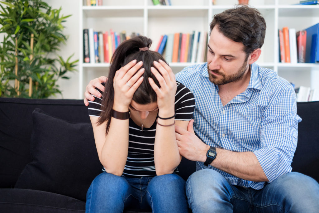 A caucasian man comforting a caucasian female as she sits with her head in her hands on a couch.