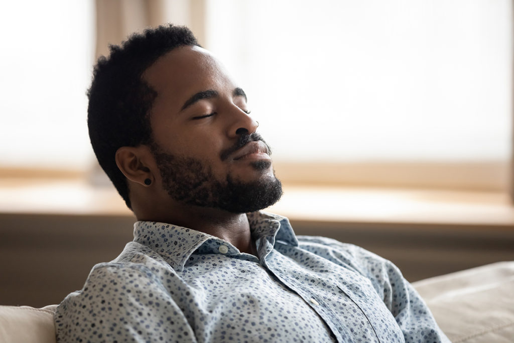 An african-american man sitting on a couch with his eyes closed. He looks peaceful.