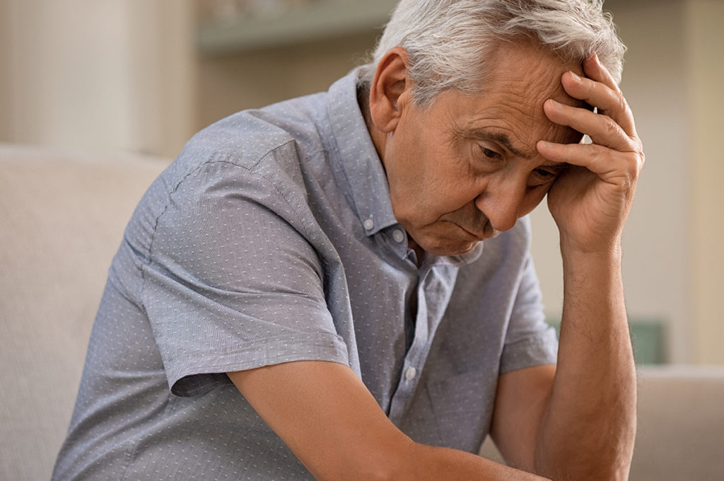 A older caucasian man sitting on a couch with his head in his hands. He appears depressed.