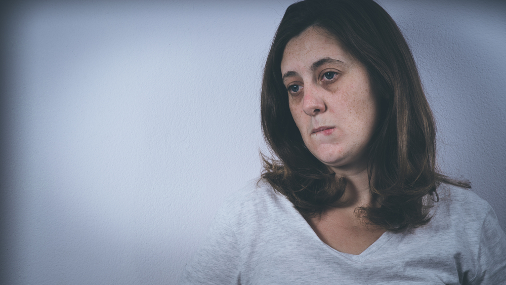 A caucasian woman sitting against a wall looking depressed.