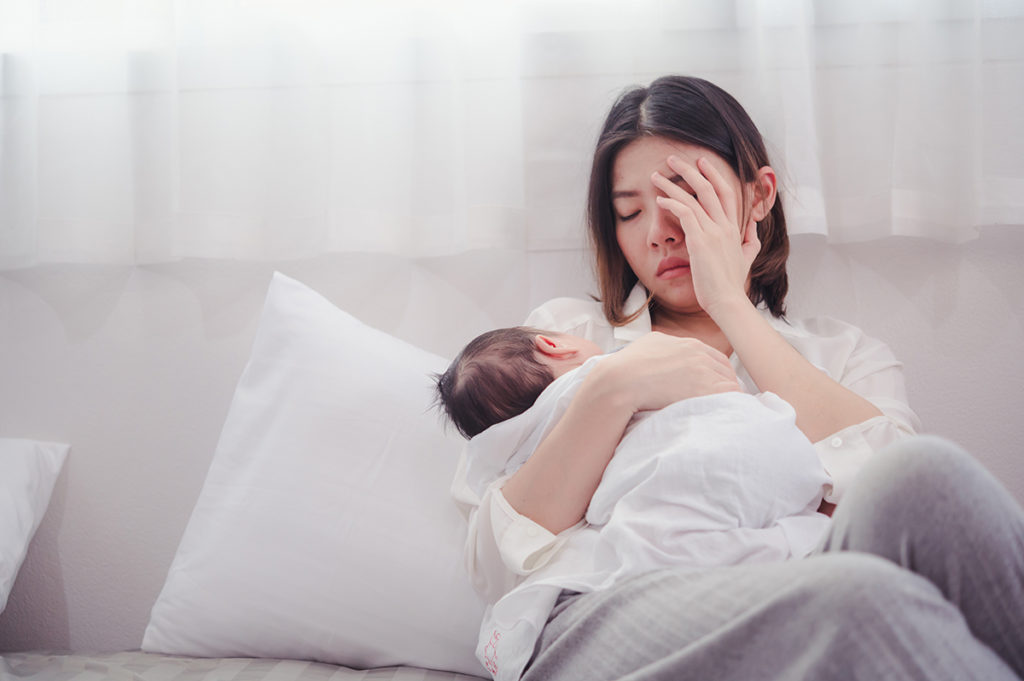 An asian mother holding a baby. She his covering her face with her hands and appears to be suffering from postpartum depression.