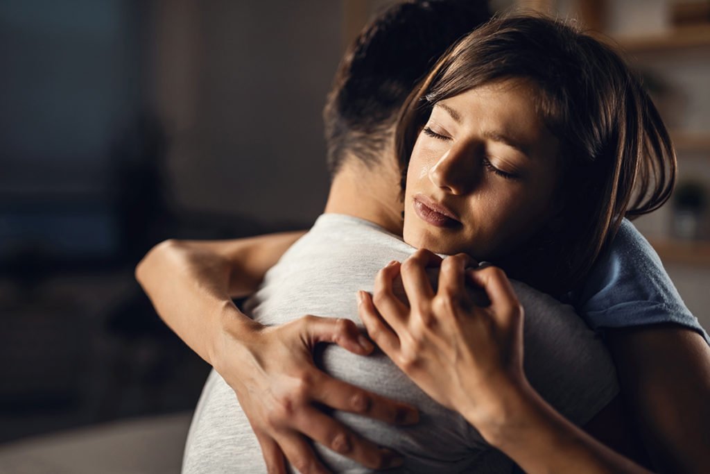 A caucasian woman hugging a caucasian male closely. She has been crying.