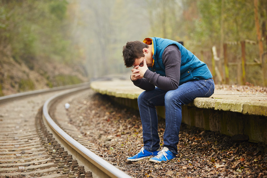 A caucasian man sitting near a rail track with his head in his hands.