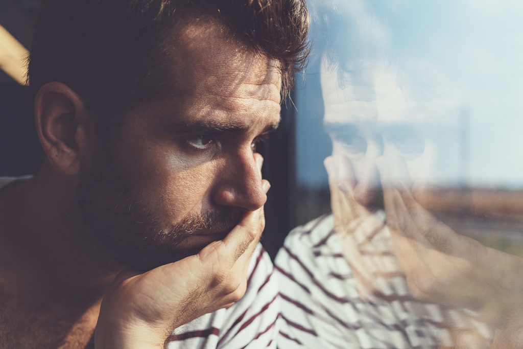 A young man with a beard looking out a window pensively with his head in his hands.