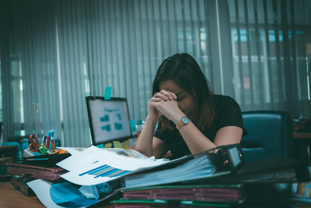 A woman sitting at her desk with her head in her hands. She is surrounded by large piles of paperwork and files.