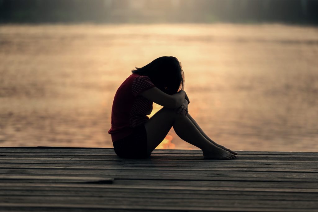 A woman sitting with her head in her hands on a deck with the water and sunset behind her.