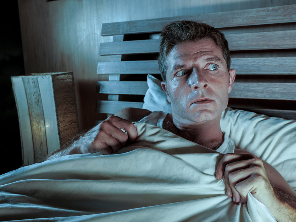 A caucasian male sitting up in bed with a fearful look on his face.