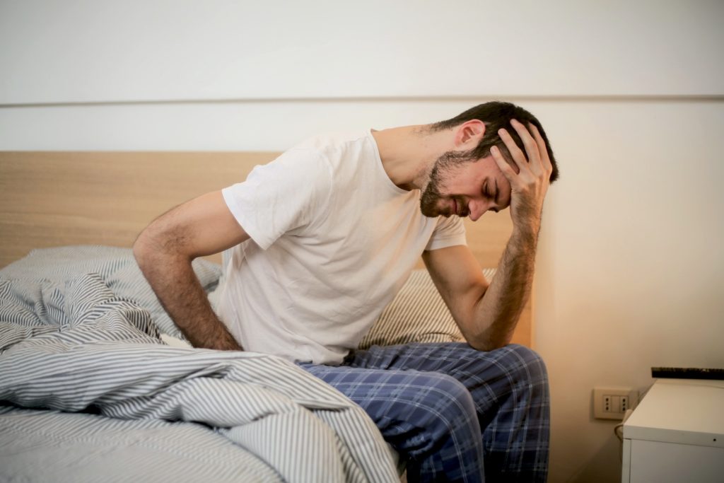 A distressed caucasian male sitting on the edge of his bed with his head in his hands.