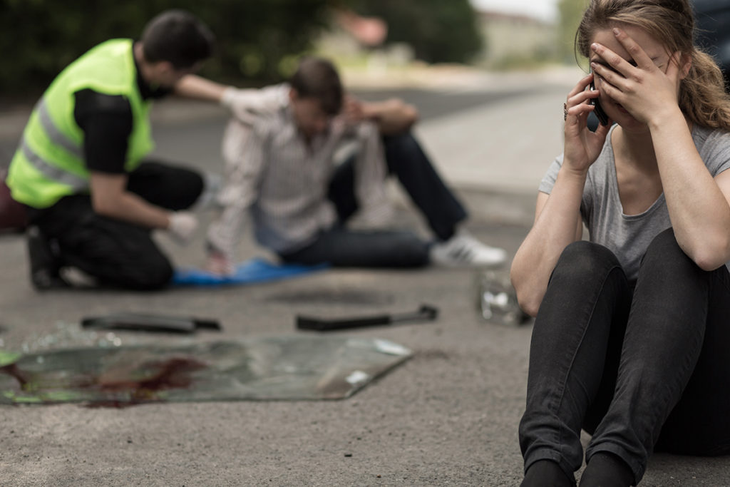A caucasian woman sitting on the road crying while on the phone. Behind her a caucasian man is being seen to by a paramedic.