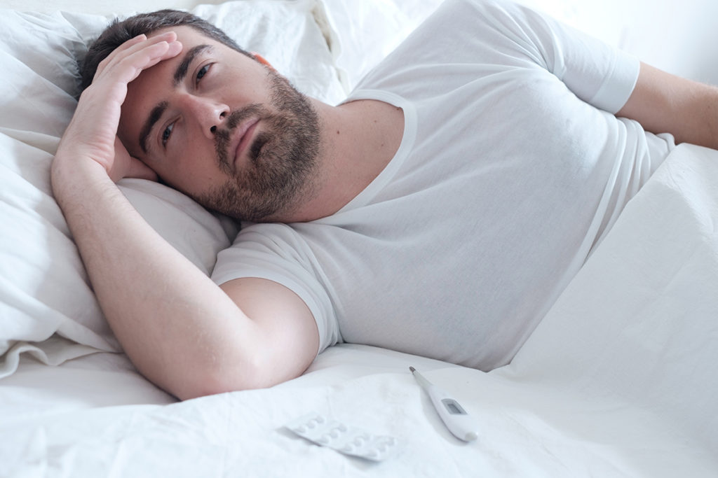 A caucasian man lying in bed with his head in his hands. He appears sick. There is a thermometer and pack of pills on the bed in front of him.