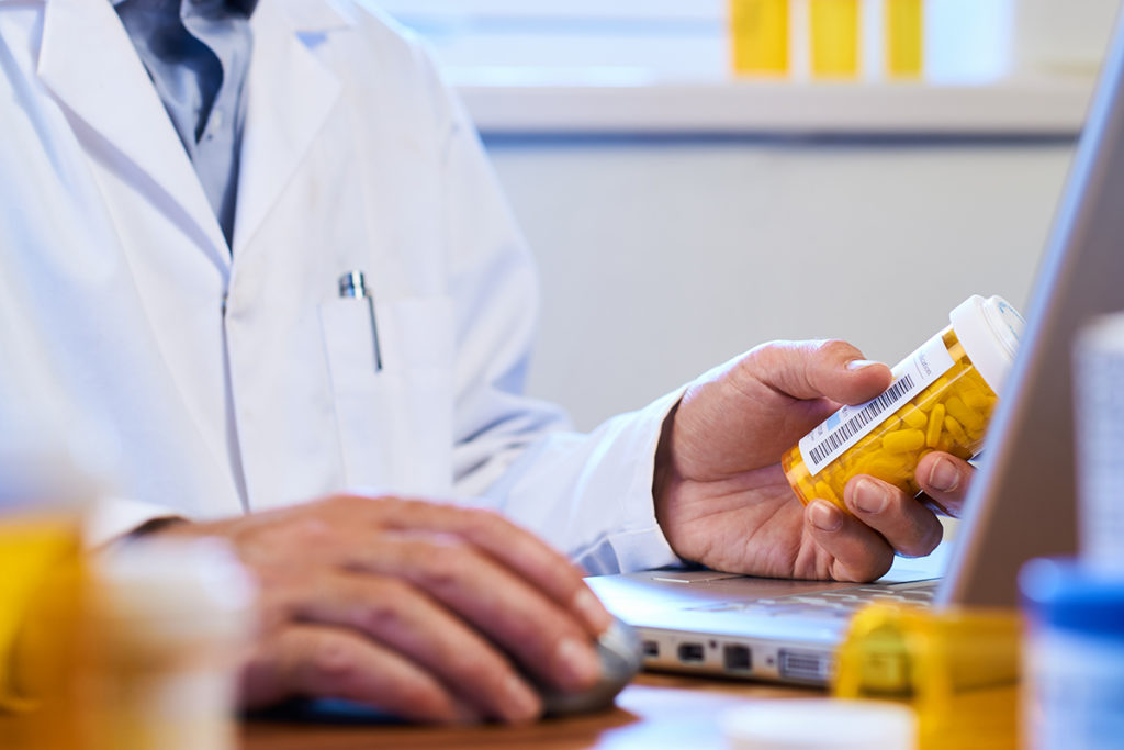 A doctor sitting in front of a laptop while holding a bottle of prescription pills