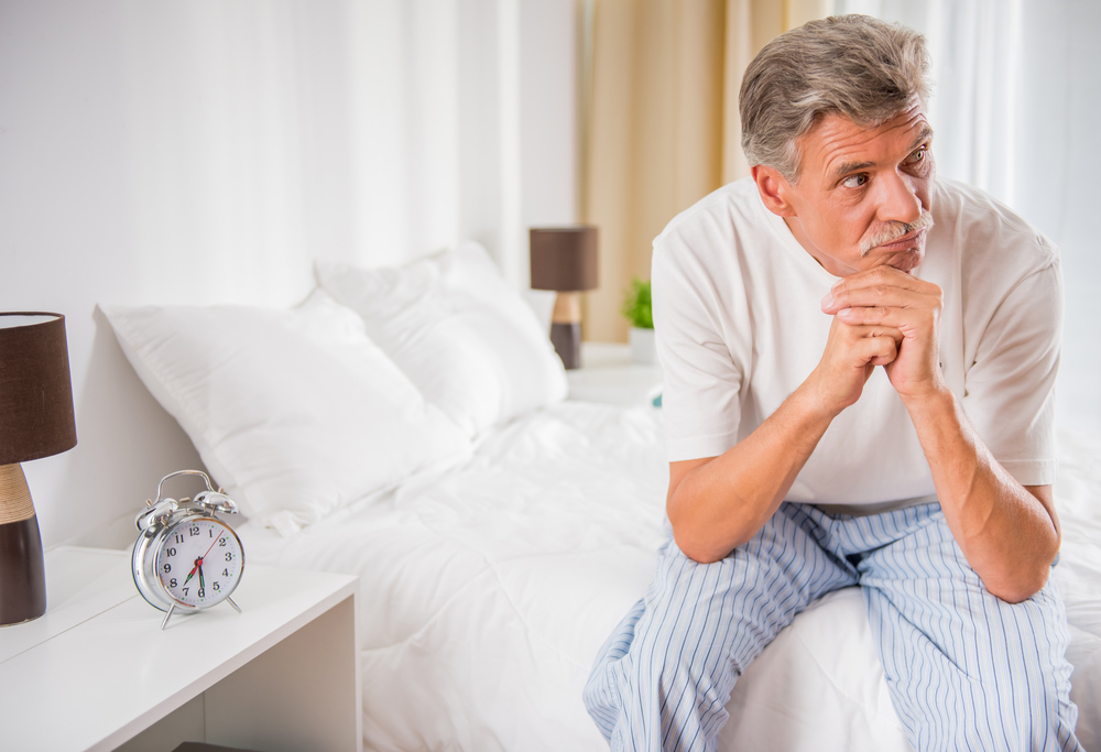 An older male sitting at the edge of his bed looking away.