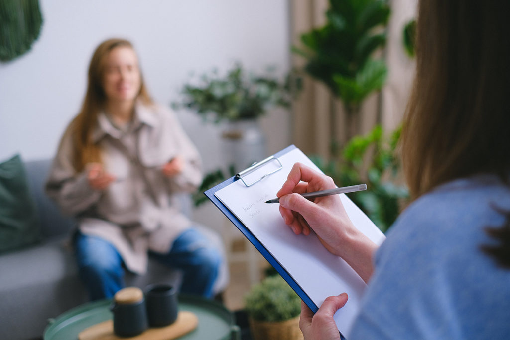 A close up of a therapy session. The therapist and her clipboard are in the forefront with her female patient in the background.