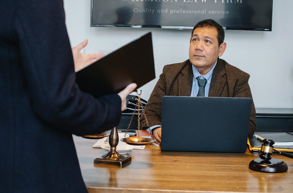 Two men having a discussion in the office. One man is standing with notes in his hand, the other is sitting with a laptop in front of him.