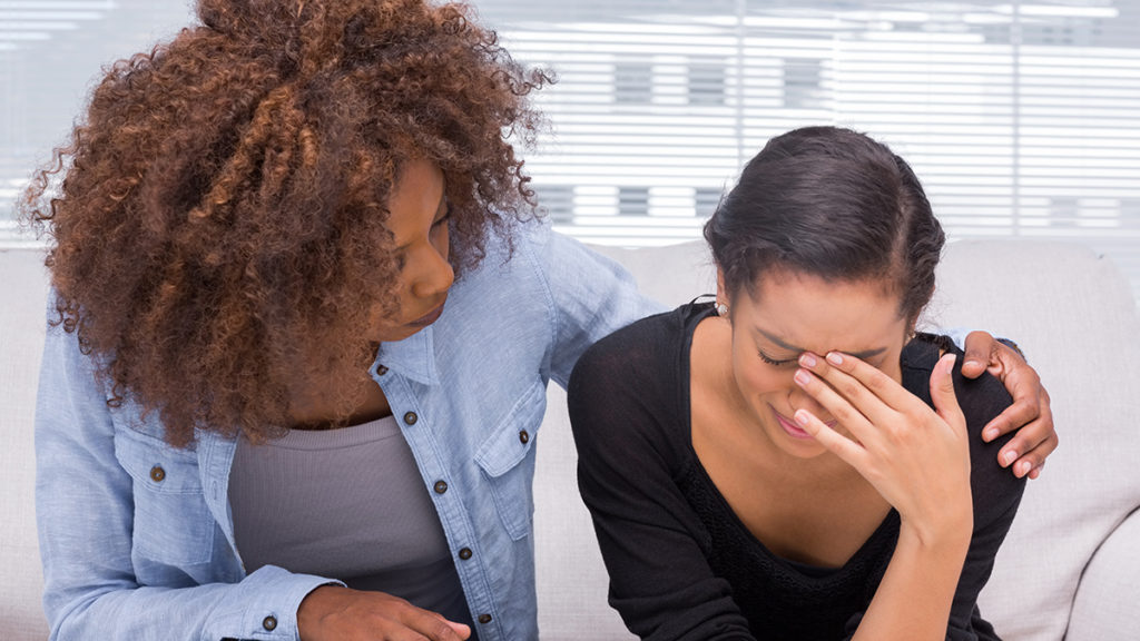 Two women sitting on a couch. One woman is comforting her crying friend.
