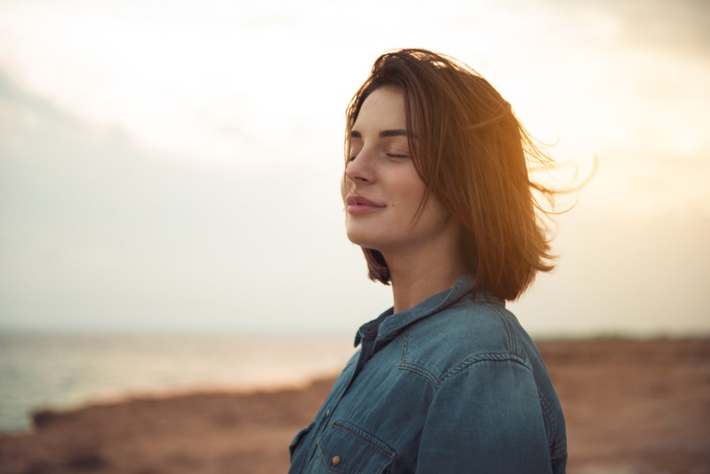 A caucasian woman smiling serenely with her eyes closed. The sun is setting behind her.