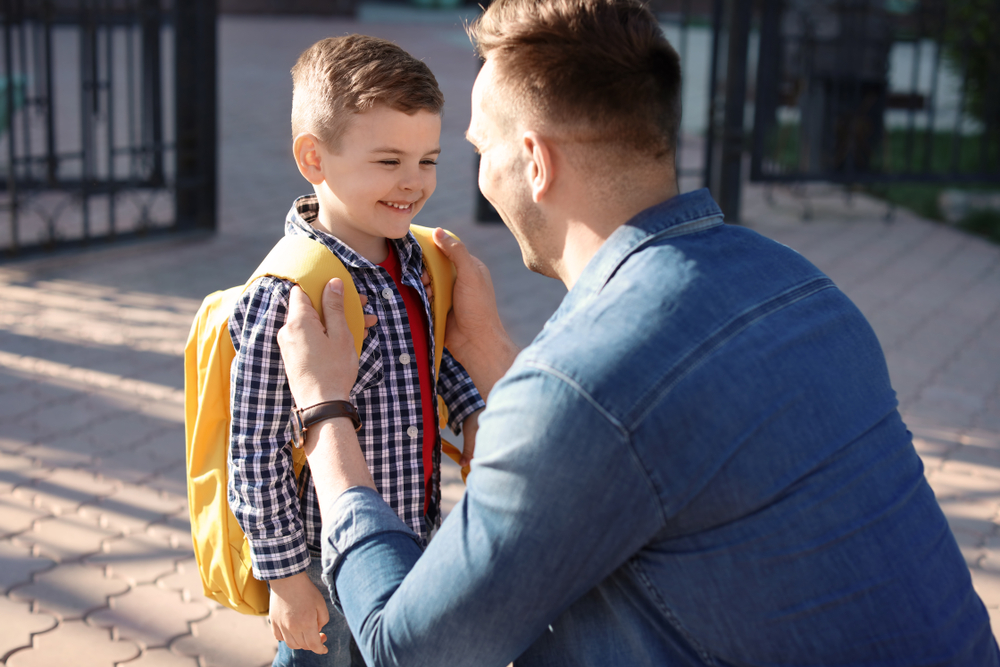 A father smiling at his son while helping him put a yellow backpack on