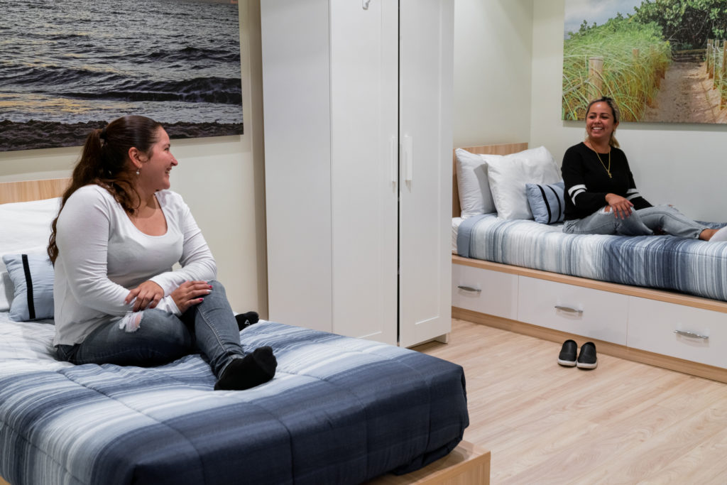 Two women sitting on their beds at Beach House Recovery center. They are smiling at each other.