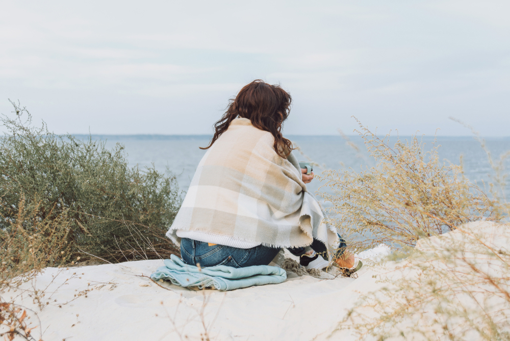 A caucasian woman sitting on the beach staring out into the ocean. She is holding a cup of coffee with a blanket around her.