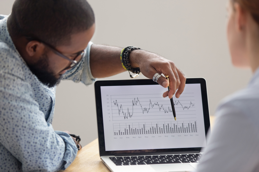 An african-american man pointing to a graph on a laptop while talking to his caucasian colleague.