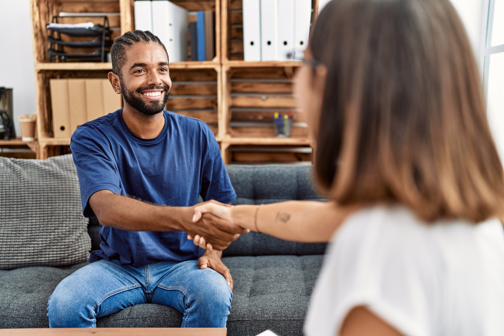 An african-american man sitting on a couch smiling at a caucasian woman and shaking her hand.