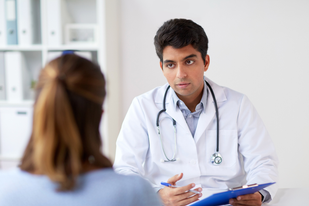 An indian doctor talking to a female caucasian patient. He is holding a clipboard and pen.