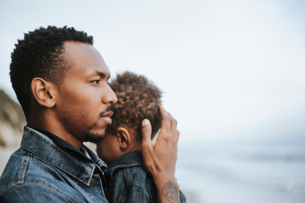 An african-american man holding his son while looking away pensively