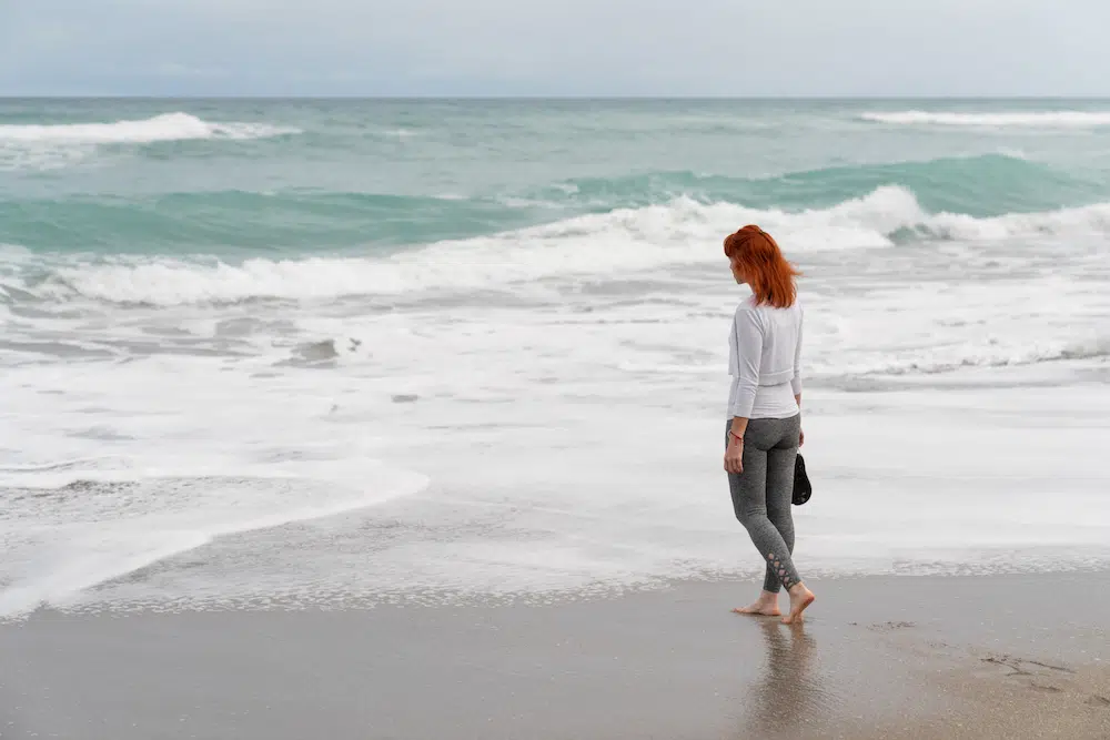 A caucasian woman walking on the beach with the waves crashing on the shore in the background