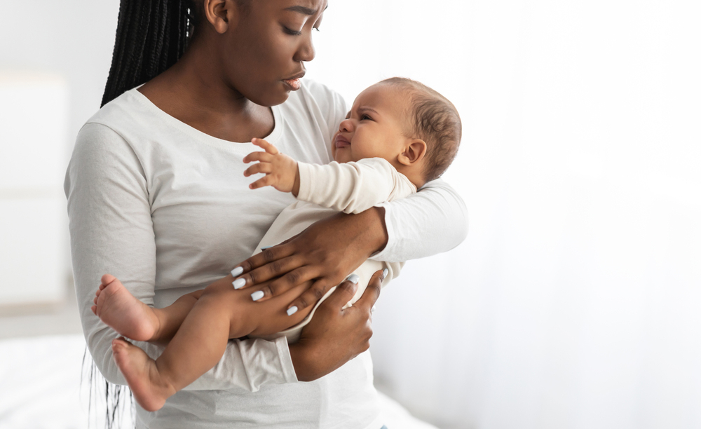 An african-american woman holding her crying baby. She looks distressed.