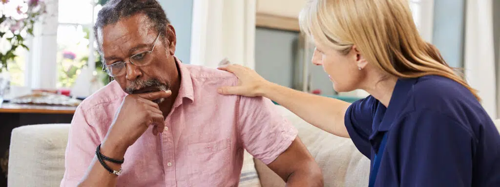 A caucasian woman sitting on a couch with a sad looking african-american man. She is comforting him.