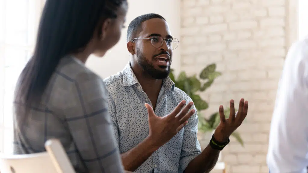 A man speaking and gesturing during a group counseling session while other participants listens.