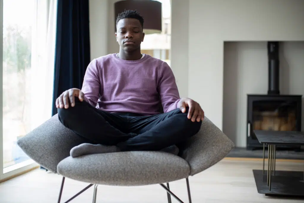 Young man with closed eyes sitting on a chair and meditating peacefully indoors