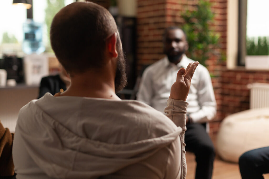 A man gesturing with his hand while speaking to a support group in a modern, casual office setting.