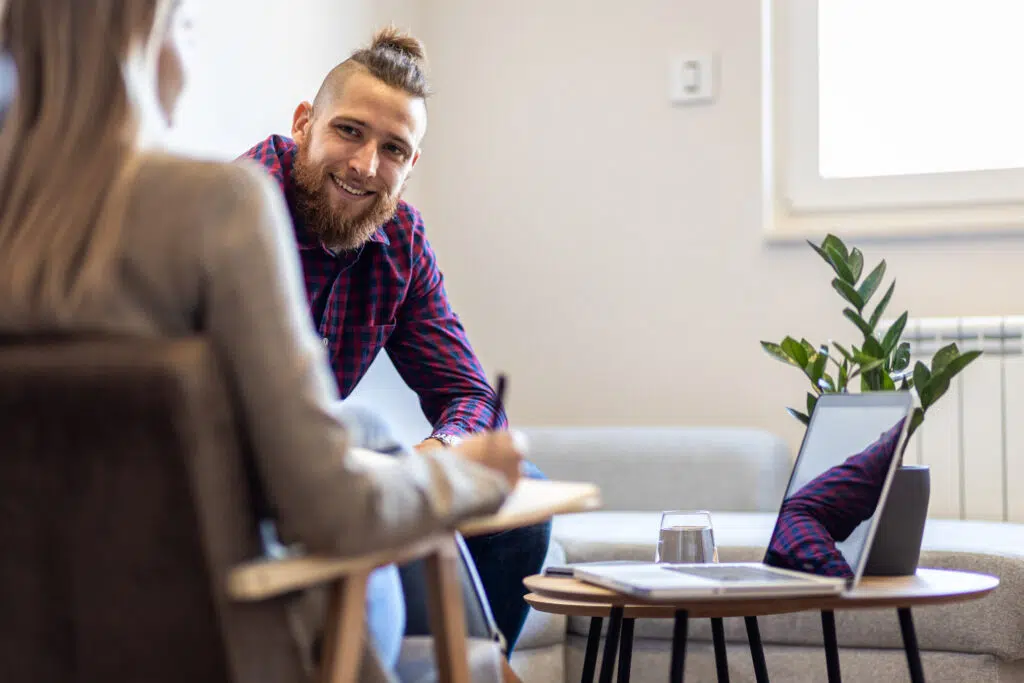 Psychologist with a clipboard listening to young male client seated on couch during a therapy planning session in modern office