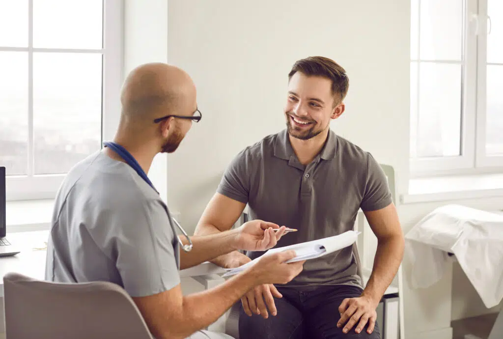 Young male patient smiling while consulting with doctor in medical office, doctor holding medical report file
