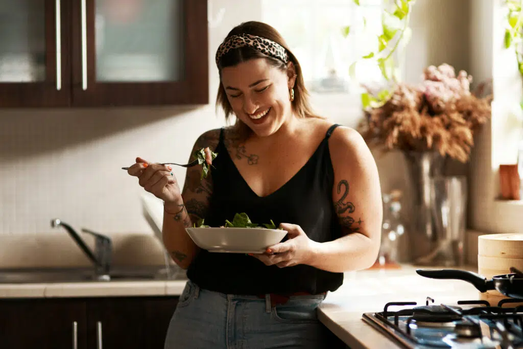 Woman smiling while eating a fresh salad with a fork in a bright kitchen setting