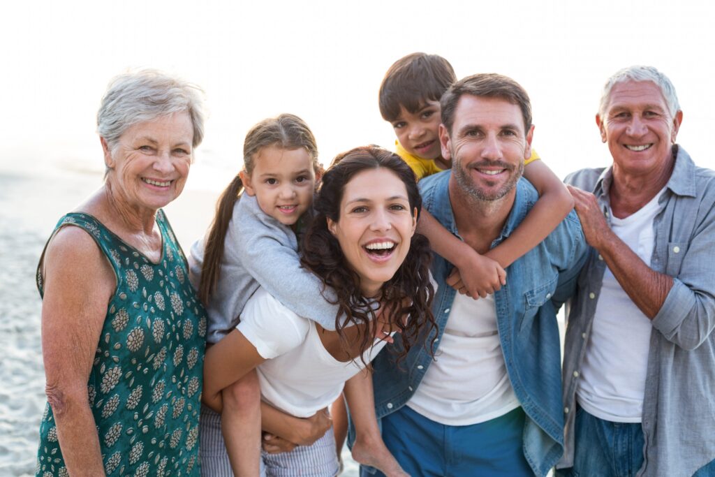 Family of two parents, two children, and two grandparents standing together on sandy beach with ocean in background, smiling at camera.
