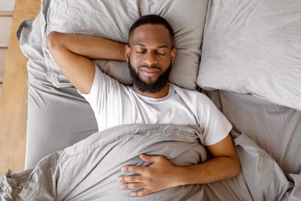 Young African American man lying on his back in bed with closed eyes, smiling peacefully while resting in morning light.