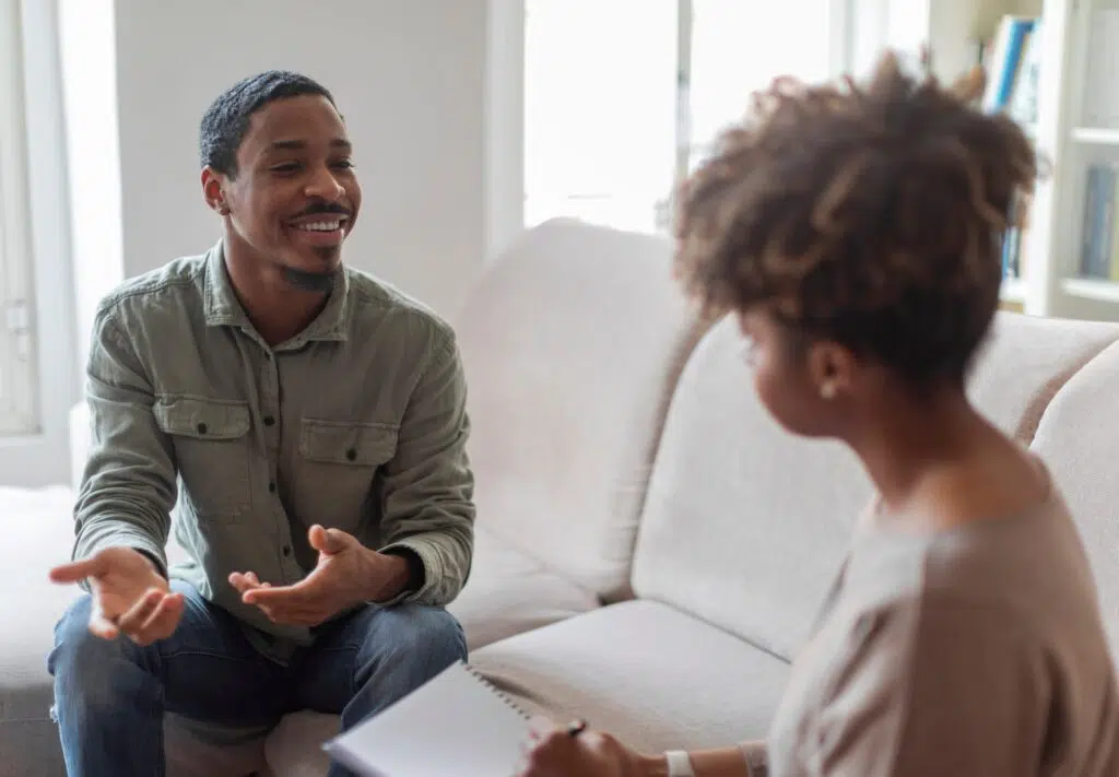 Young African American man sitting on couch in therapy session, smiling while talking with female therapist in counseling office.